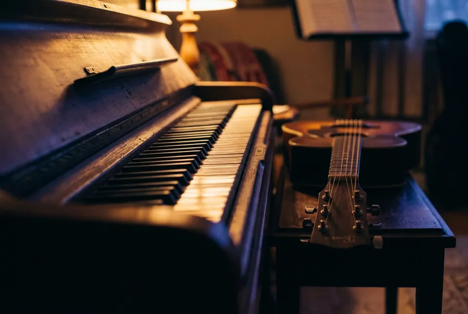 Le clavier d'un piano droit vu de près, une guitare posée sur un banc à côté, sous la lumière d'une lampe.